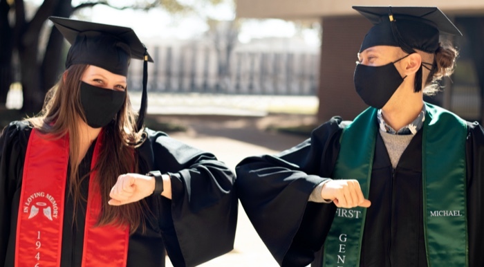 Two graduates wearing black caps, gowns, masks and stoles bumping elbows. Red stole on left grad, green stole on right grad.