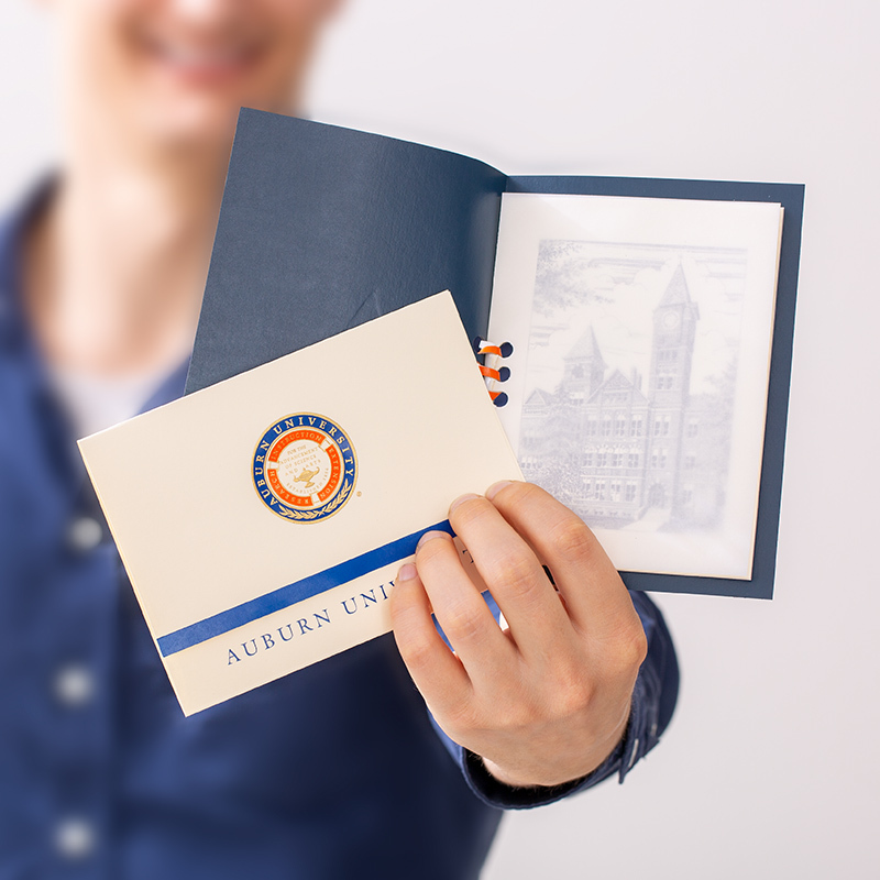 Hands holding a college announcement in blue and orange foil, along with a blue keepsake announcement holder.