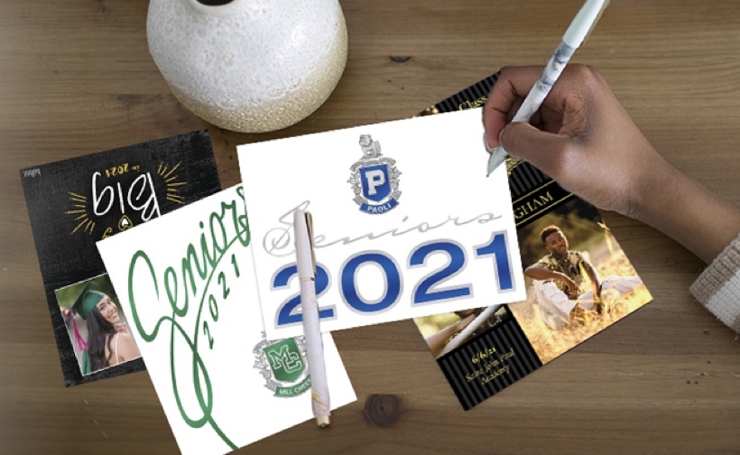 Four Class of 2021 graduation announcement designs lying on a wood desk. A hand is writing on one announcement with a pen.